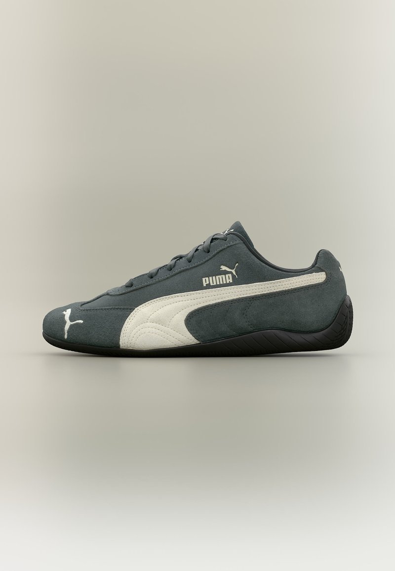 Σκούρο γκρι αθλητικό παπούτσι Puma με λευκή ρίγα στο πλάι, λογότυπο Puma στο πλάι και στα δάχτυλα, και μαύρη υφασμάτινη σόλα, παρουσιάζεται σε προφίλ σε ουδέτερο φόντο.
