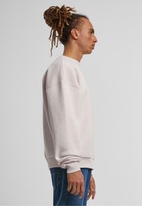 Lys rosa bomullssweater med avslappet passform, senkede skuldre og ribbede mansjetter. Brukt over blå jeans, sett fra siden.