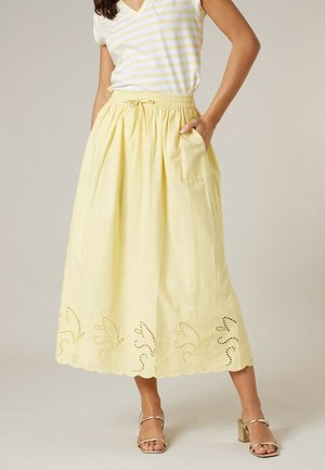 Femme portant une jupe midi jaune avec un bord en broderie œillet, de grandes poches, une taille à cordon, associée à un haut rayé jaune et blanc et des talons dorés.