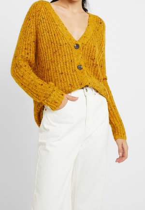 Personne portant un cardigan en maille épaisse jaune moutarde avec deux boutons et un pantalon blanc taille haute, main dans la poche, sur un fond uni.