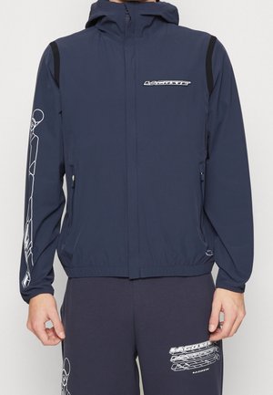 Homme portant une veste à capuche bleu marine et un pantalon assorti avec des imprimés graphiques blancs et des logos Lacoste sur la poitrine, le bras et la jambe.