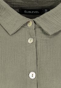 Olive green button-up shirt with a textured fabric, featuring a pointed collar and subtle stitching. Pearl-like buttons on the placket.