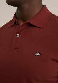 Tricou polo bordo realizat dintr-un material texturat, cu o închidere cu două butoane, prezentând un logo brodat cu o pasăre pe partea stângă a pieptului.