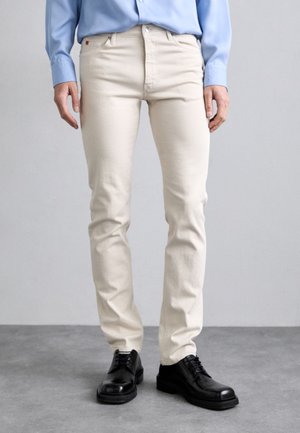 Beige cotton jeans with a slim fit, five-pocket design, and metal rivets, paired with black leather shoes. Smooth texture, casual style.