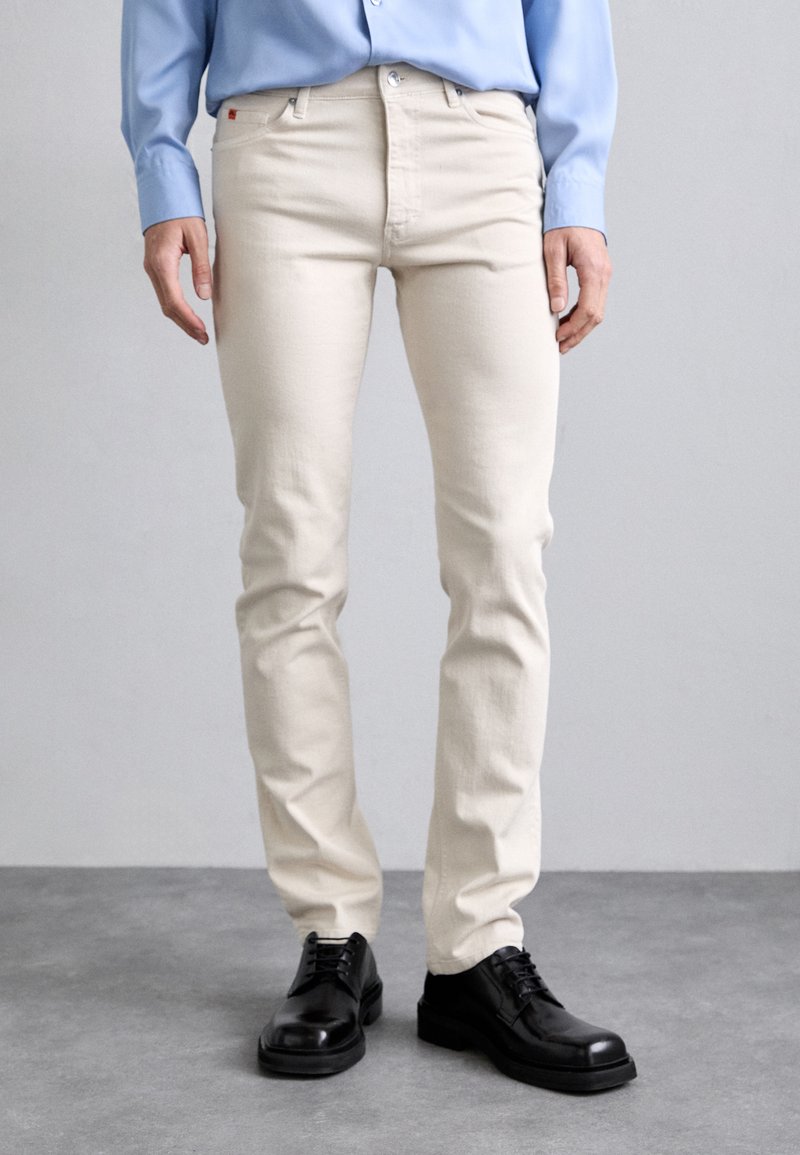 Beige cotton jeans with a slim fit, five-pocket design, and metal rivets, paired with black leather shoes. Smooth texture, casual style.