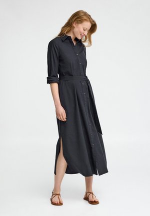 Mujer con cabello rojo que lleva un vestido camisero negro con botones, cinturón, aberturas laterales y sandalias marrones, mirando hacia abajo y sonriendo.