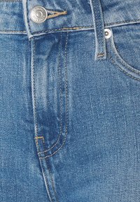 Blå denimjeans med en texturerad yta, som har en framficka och en metallknappstängning med varumärkeslogotypen synlig.
