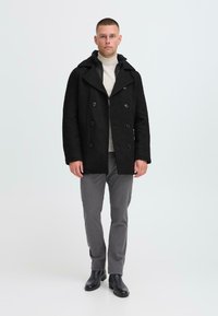 Schwarzer doppelreihiger Wollmantel mit großen Knöpfen, breiten Revers und Kapuze, getragen über einem hellen Rollkragenpullover und grauen Hosen.