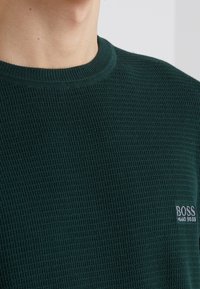 Ciemnozielony sweter o teksturowanej strukturze z okrągłym dekoltem i małym haftowanym logo "BOSS" po lewej stronie. Normalny krój i tkany materiał.
