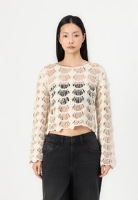 Pulôver cropped em estilo de crochê em bege claro, com um padrão de conchas aberto e mangas longas. Combinado com calças pretas de cintura alta.