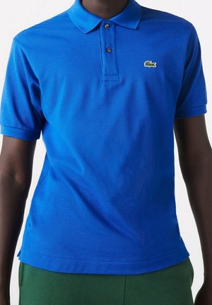 Person trägt ein hellblaues kurzärmeliges Poloshirt mit kleinem grünem Krokodil-Logo auf der Brust, kombiniert mit grüner Hose.