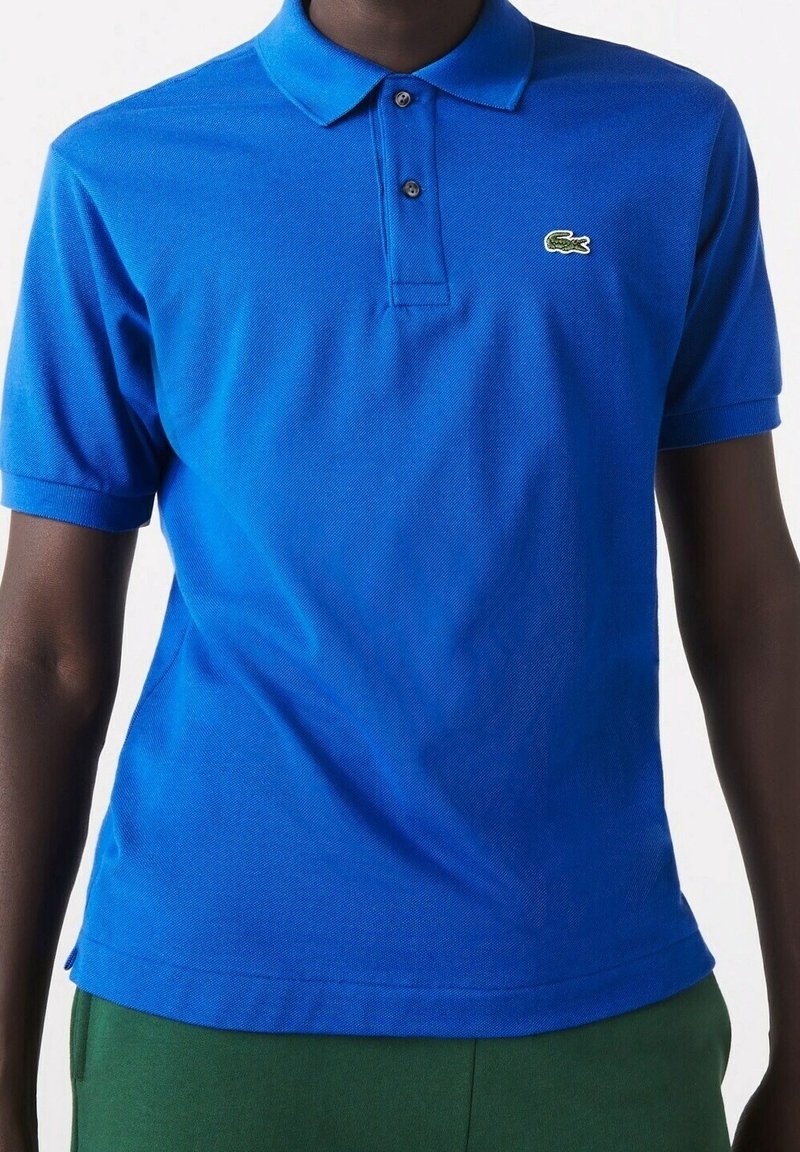 Personne portant un polo bleu vif à manches courtes avec un petit logo crocodile vert sur la poitrine, assorti à un pantalon vert.