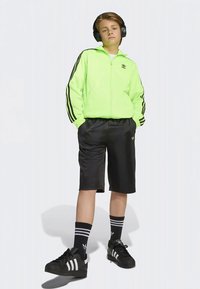 Neon-grüne Zip-Jacke mit schwarzen Streifen, schwarze Shorts und schwarze Schuhe. Drei Streifen auf den Ärmeln und Socken, das Adidas-Logo ist sichtbar.