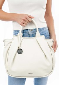 Bolso de cuero blanco con acabado texturizado, que cuenta con una cremallera, una etiqueta de metal y dos asas, sostenido por una persona que lleva vaqueros de color azul claro.