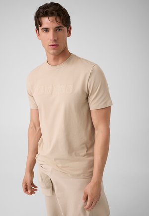 Jeune homme aux cheveux courts et bouclés portant un t-shirt Guess beige et un pantalon beige assorti, debout devant un fond gris uni.