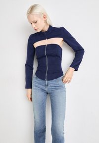 Navy blue fabric top with a high collar, asymmetrical cutout, and front zipper. Long sleeves, fitted design, and textured material.