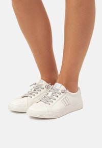 Zapatillas blancas con cordones grises, punta redonda y acabado en cuero texturizado. Presentan un logo sutil en el lado y suela de goma plana.