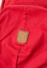 Κόκκινη υφασμάτινη τσάντα πλάτης που δείχνει μια ραμμένη τσέπη με μια καφέ δερμάτινη επένδυση που διαθέτει ένα έμβλημα αλεπούς και έναν κυκλικό σχεδιασμό.