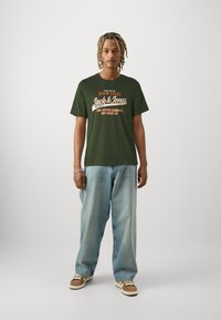 Olive green cotton t-shirt with white and orange printed text, round neckline, paired with light blue wide-leg jeans and beige sneakers.