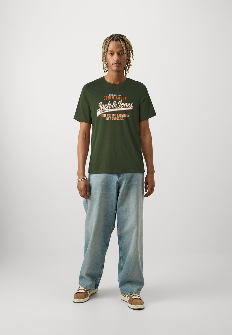 Olive green cotton t-shirt with white and orange printed text, round neckline, paired with light blue wide-leg jeans and beige sneakers.