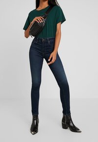 Mörkblå skinny jeans med hög midja, i kombination med en grön t-shirt med kort ärm och en svart axelväska. Svarta ankelstövlar med spetsig tå.