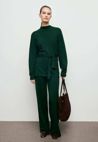 Grünes Strickoberteil mit hohem Kragen und tailliertem Bund, kombiniert mit weiten grünen Strickhosen. Das Model hält eine braune Tragetasche und trägt braune Schuhe.