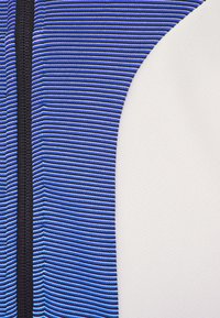 Blåt og sort stribet tekstil integreret med et glat hvidt stof, der har et tekstureret mønster på den hvide sektion og en lynlås.