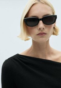 Schwarze Cat-Eye-Sonnenbrille mit dunklen Gläsern. Modell trägt ein schwarzes Off-Shoulder-Top. Glatte Texturen und moderne Designelemente sind deutlich erkennbar.