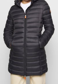 Schwarze Pufferjacke mit horizontalem Steppmuster, einem Reißverschluss vorne und einer verstellbaren Kapuze. Besitzt orangefarbene Hardware-Details und seitliche Taschen.