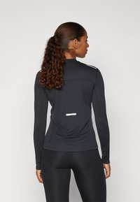 Schwarzes, langärmeliges Sportoberteil mit einem Stehkragen, ausgestattet mit reflektierenden Akzenten und einem strukturierten Stoff. Schmeichelhafte Passform über einer schwarzen Leggings.