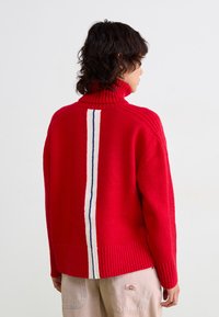 Rode coltrui met ribbeltextuur, voorzien van een verticale streep in wit en blauw langs de achterkant. Gebreide stof, casual pasvorm.