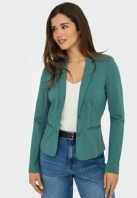 Veste bleu-vert en tissu lisse avec une coupe ajustée, fermeture à un bouton et deux poches avant, associée à un top blanc et un jean bleu.