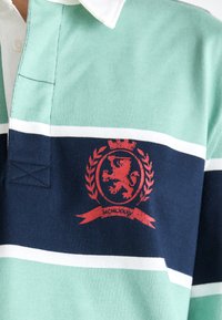Nærbilde av en stripete polo-skjorte med mintgrønne, marineblå og hvite striper, med et rødt løveemblem på brystet.