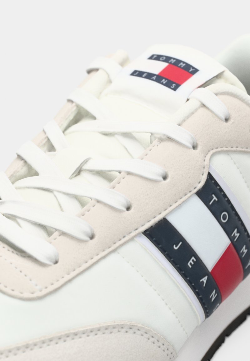 Zapatilla blanca y beige con material de ante, cierre de cordones y un logo en el lateral que presenta franjas azules, rojas y blancas.