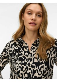 Femme aux cheveux châtain clair ondulés portant des boucles d'oreilles argentées en forme d'anneau et un chemisier boutonné à motifs noir et blanc, sur un fond blanc uni.