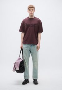Vinrød oversized T-shirt med præget "HUGO" logo, lyseblå jeans, sorte sko og en sort taske med en lilla sweater ovenpå.