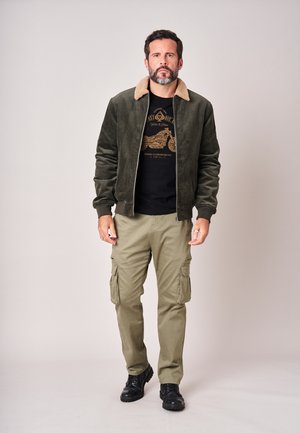 Man met baard die een jas met groene shearlingkraag draagt, een zwart grafisch t-shirt, beige cargobroeken en zwarte laarzen, staand tegen een effen achtergrond.