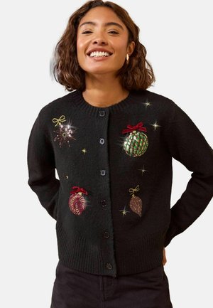Frau lächelt, trägt einen schwarzen, geknöpften Cardigan, der mit glitzernden Weihnachtsornamenten und roten Schleifen auf einem einfarbigen Hintergrund dekoriert ist.