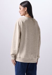 Beige sweatshirt met een ronde halslijn, ribbel-mouwen en -zoom, losse pasvorm, gedragen met blauwe spijkerbroek. Vlotte textuur, casual ontwerp.