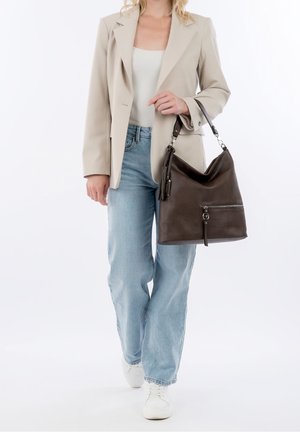 Femme portant un blazer beige, un haut blanc, un jean bleu clair, des baskets blanches, et tenant un grand sac à main en cuir marron foncé sur l'avant-bras.