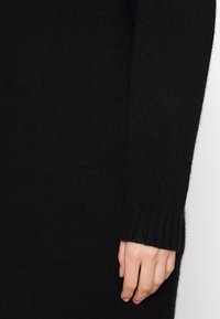 Maglione nero a maglia con texture, caratterizzato da un polsino a costine e un primo piano di una mano che tiene la manica. Tessuto morbido e di peso medio.