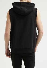 Felpa senza maniche nera con tasca frontale, cappuccio con cordini e dettagli cuciti. Tessuto morbido con una texture liscia e vestibilità casual.