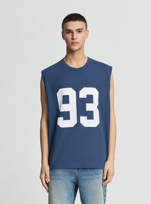Młody mężczyzna ubrany w granatowy bezrękawnik z dużym białym nadrukiem "93", w połączeniu ze jasnoniebieskimi jeansami, stoi na jednolitym tle.