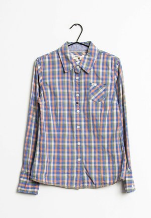 Langærmet skjorte med knapper og tern i blå, orange og grøn, hængende på en sort bøjle mod en hvid baggrund.