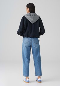 Sweatshirt noir avec capuche grise, ourlet élastiqué, associé à un jean droit bleu clair et des baskets rayées bleues, vu de dos.