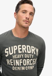 Grijze longsleeve t-shirt met een grote, getextureerde print in crème met de tekst "SUPERDRY HEAVY DUTY REINFORCED DENIM COMP." Ronde halslijn.