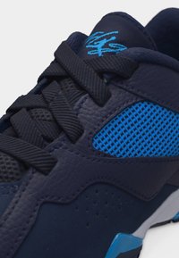 Κοντινό πλάνο σε σκούρο μπλε αθλητικό παπούτσι με μαύρα κορδόνια, μπλε πλέγμα πλαϊνό πάνελ, υφή δέρματος και ανοιχτό μπλε λογότυπο στη μαλακή γλώσσα.