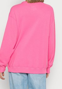 Jasnoróżowy, oversize'owy sweatshirt z okrągłym dekoltem, ściągaczami przy rękawach i u dołu. Gładka tekstura, luźny krój. Noszony z jasnoniebieskimi dżinsami.
