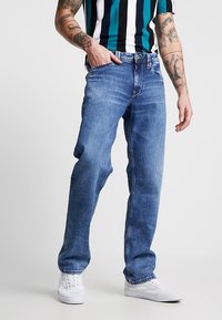 Vaqueros azules de denim con un ajuste relajado, con pierna recta, diseño clásico de cinco bolsillos y un sutil desgaste, combinados con zapatillas blancas.