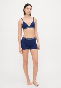 Navy modra spodnja perila sestava, ki vključuje brezžični modrček z globokim V izrezom in ujemajoče se spodnje hlačke boyshorts. Obe kosi imata elastičen trak z logotipom.
